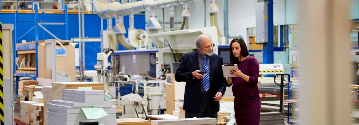 Two professionals review a tablet on a factory floor.