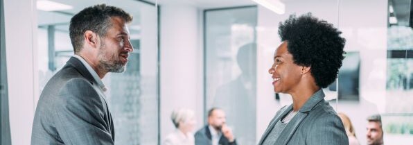 Due professionisti in abiti formali si stringono la mano durante un incontro di lavoro in una sala riunioni moderna, con altri colleghi sullo sfondo.