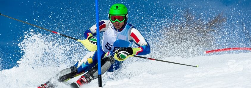 Scialpinista in gara mentre affronta una curva tra i pali su una pista innevata, sollevando una nuvola di neve durante la discesa.