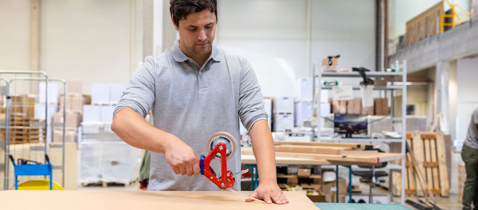 Worker sealing a cardboard box with a tape gun in a warehouse, surrounded by stacked packaging materials and pallets.