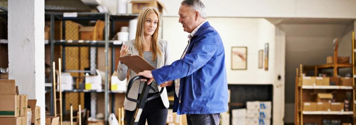 Two colleagues discuss inventory using a clipboard in a warehouse.