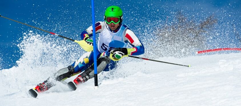 Scialpinista in gara mentre affronta una curva tra i pali su una pista innevata, sollevando una nuvola di neve durante la discesa.