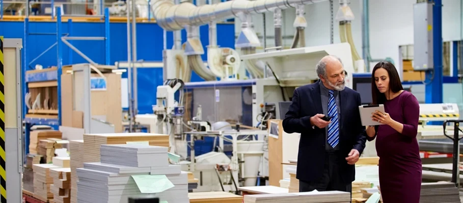 Two professionals review a tablet on a factory floor.