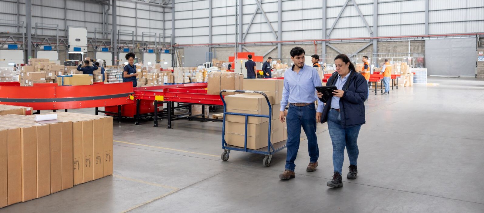 Two colleagues reviewing a tablet while walking through a large distribution warehouse with conveyor belts and packed boxes in the background.