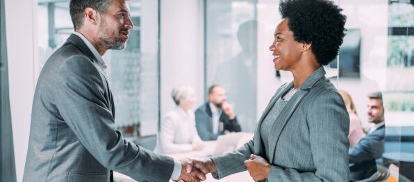 Due professionisti in abiti formali si stringono la mano durante un incontro di lavoro in una sala riunioni moderna, con altri colleghi sullo sfondo.